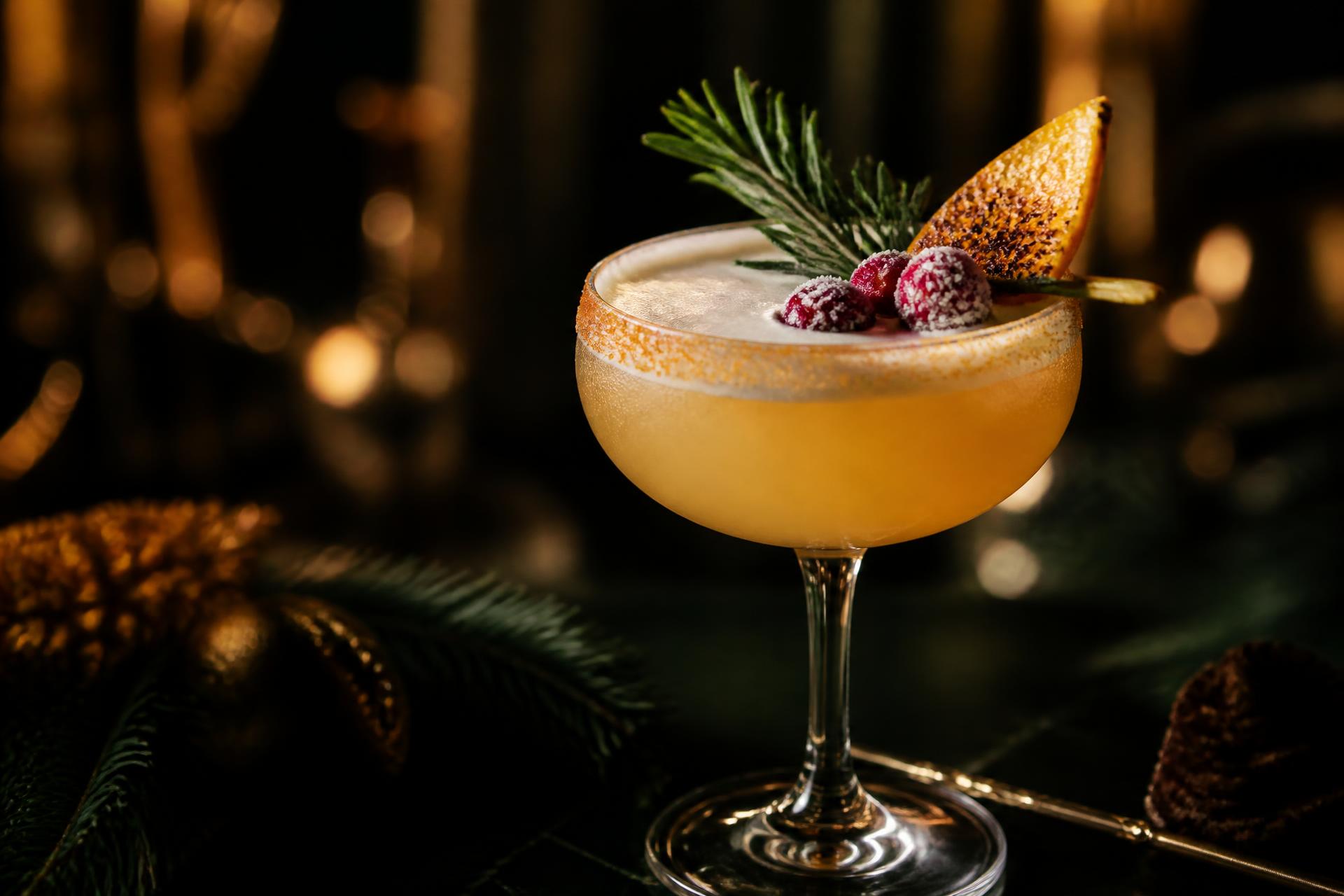 Holiday cocktail in a coupe glass garnished with rosemary, sugared cranberries and torched orange